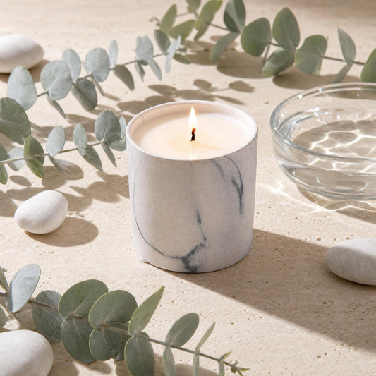 Marble-look white candle in ceramic jar with lit wick on light stone surface